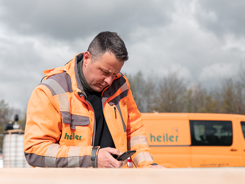 Sebastian ist Polier und steht vor einem organgenen Bulli und hält sein Telefon in der Hand